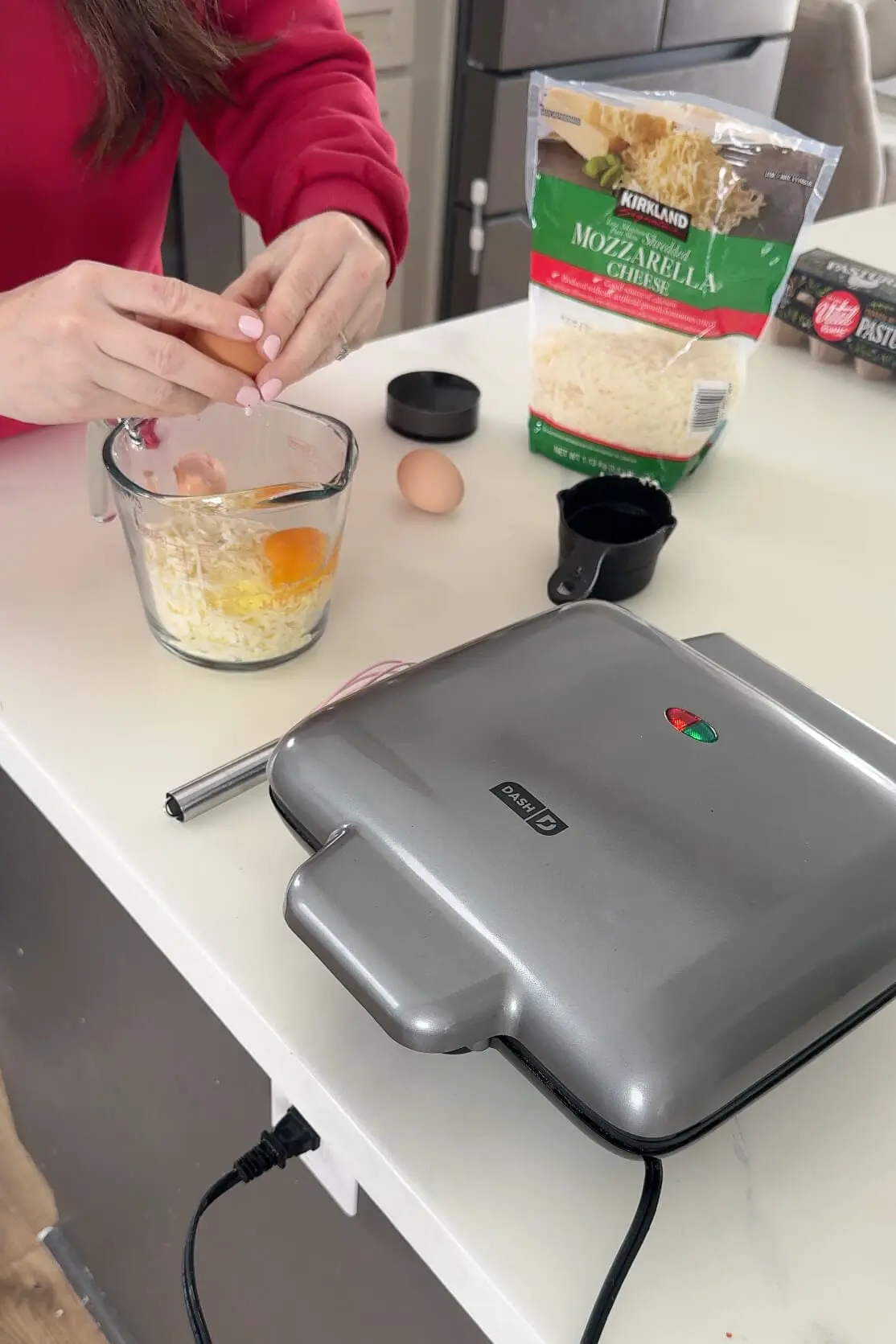 A woman in a red sweater cracks a fresh egg into a measuring cup filled with shredded mozzarella cheese. A bag of cheese, a measuring cup, and a waffle maker are visible on the counter.